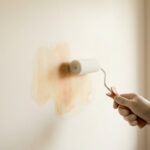 Une main gantée utilisant un rouleau de peinture sur un mur beige présentant une grande tache jaune.